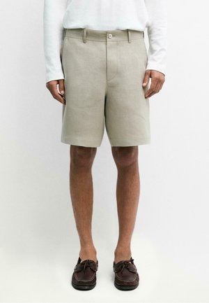 Mann, der beige knielange Shorts, ein weißes langärmliges Hemd und dunkelbraune Mokassins trägt, steht vor einem schlichten weißen Hintergrund.