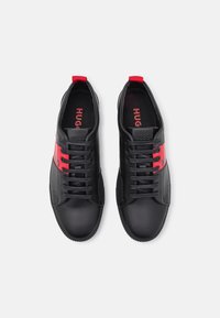 Černé kožené tenisky mají červený akcentní pás, ploché tkaničky a texturovanou gumovou podrážku. Značení je viditelné na jazyku a stranách.