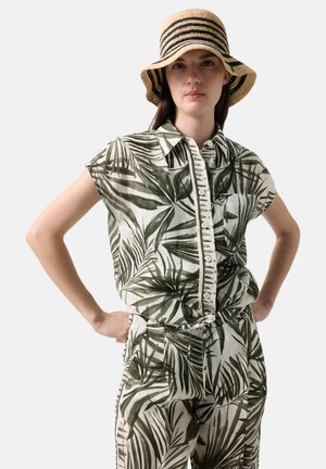 Kobieta ubrana w pasującą koszulę z guzikami i spodnie z tropikalnym liściastym wzorem w zielono-białych kolorach, z paskowanym słomkowym kapeluszem, z rękami na biodrach.