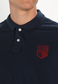 Polo din bumbac de culoare bleumarin, realizat dintr-un material texturat, având un guler cu trei nasturi și un logo brodat în roșu cu stele și un design în formă de scut.