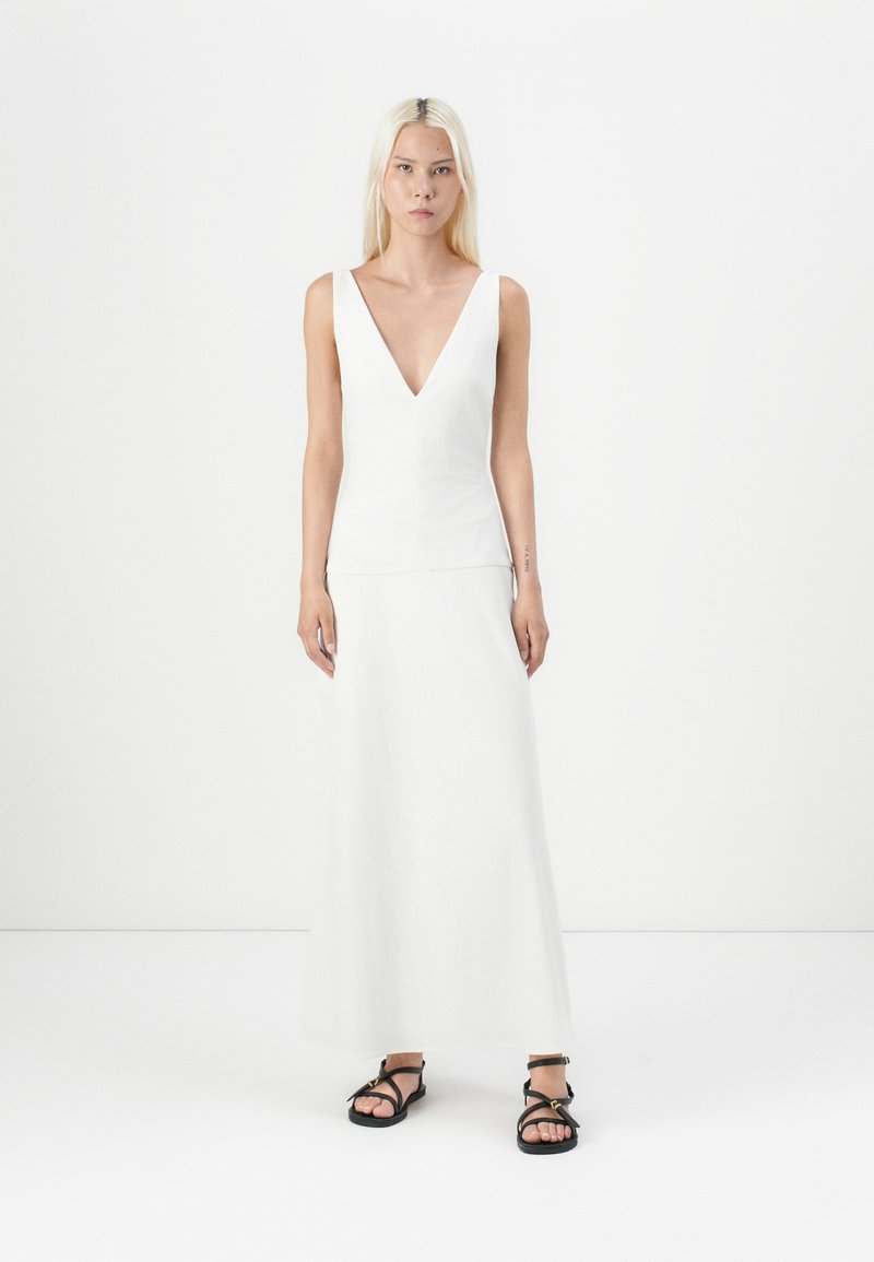 Vestido branco sem mangas com decote em V e corpete ajustado, fluindo para uma saia longa. Combinado com sandálias pretas de tiras. Textura de tecido suave.