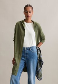 Cardigan long vert olive avec capuche, porté sur un t-shirt blanc, associé à un jean bleu. Une écharpe à motifs est glissée dans la taille.