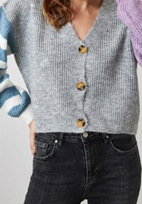 Grau gestrickte Strickjacke mit V-Ausschnitt, ausgestattet mit drei goldenen Knöpfen und gestreiften blau-weißen Ärmeln, kombiniert mit schwarzer Jeans.