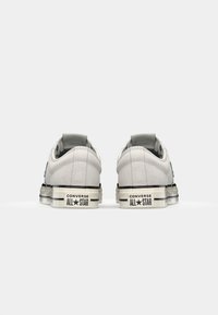 White suede sneakers with a black rubber sole, featuring a flat heel, padded collar, and Converse All Star branding on the back.