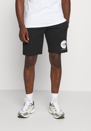 Svarta shorts i bomull med en cirkulär logotypapplikation på vänstra låret, ihop med vita strumpor och löparskor med silver- och gula detaljer.