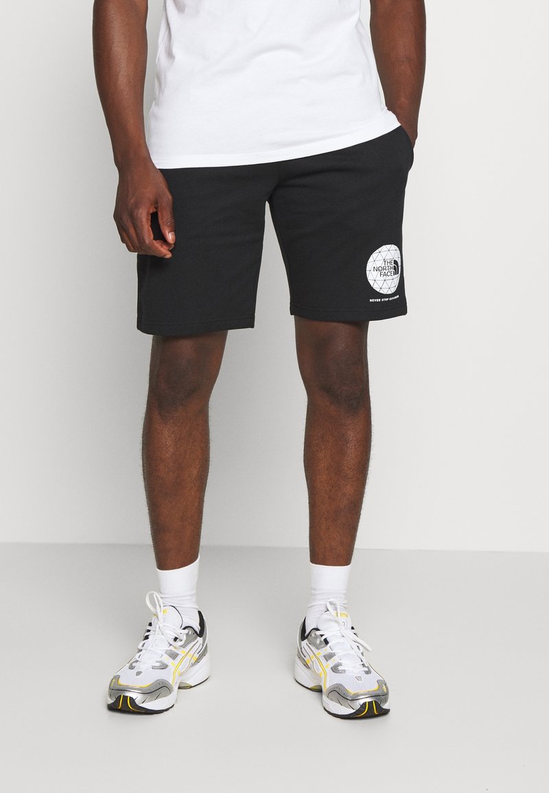 Černé šortky z bavlny, s kulatou nášivkou na levém stehně, spárované s bílými ponožkami a běžeckými botami se stříbrnými a žlutými detaily.