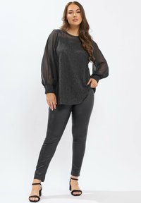 Femme portant un haut long en dentelle noire transparente avec un panneau avant en dentelle, un pantalon ajusté noir et des escarpins à bride de cheville noirs, se tenant sur un fond blanc.