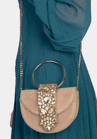 Borsa a tracolla in camoscio beige con catena metallica. Presenta una forma semicircolare e una patta frontale adornata con grandi gemme scintillanti.