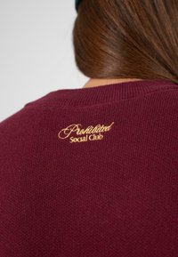 Bordowy sweter dzianinowy z haftowanym na złoto logo "Prohibited Social Club" na plecach przy dekolcie. Miękka faktura i krótki dekolt.