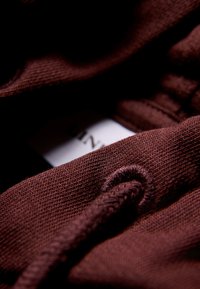 Tessuto marrone con texture, con un cordoncino visibile e un'etichetta bianca parzialmente in mostra. Il materiale sembra morbido e intrecciato.