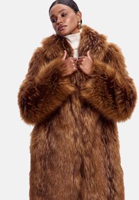 Brun faux fur frakke med lange, fluffy ærmer og en bred krave. Har en tekstureret overflade og en løs, oversized pasform.