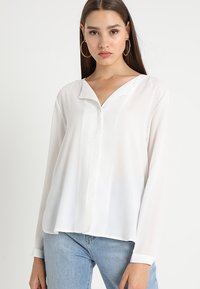 White, long-sleeve blouse with a deep V-neckline and a smooth, lightweight texture. Paired with light blue jeans.