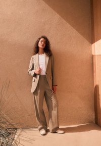 Completo beige su misura con pantaloni a gamba larga e camicia bianca aderente. La modella è in piedi contro un muro strutturato, proiettando un'ombra morbida.