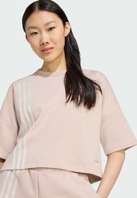 Svijetlo ružičasti skraćeni majica od mekog materijala, s tri vertikalne bijele pruge s jedne strane i malim logom na rubu.