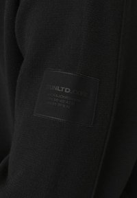 Sweatshirt noir en tissu texturé ; comporte une étiquette cousue avec "UNLTD CORE" et des détails de la marque sur la manche gauche.