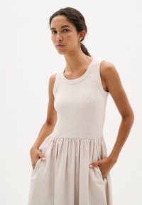 Donna che indossa un vestito beige senza maniche con corpino a coste e gonna arricciata, in piedi con le mani nelle tasche davanti a uno sfondo semplice.