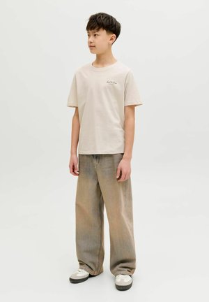 Teismeline poiss seisab, seljas helebeež lühikeste varrukatega t-särk, laiad pleekinud teksad ja valged tossud, vaatab küljele tavalise tausta ees.