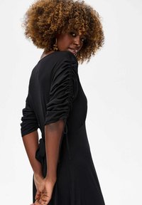 Femme aux cheveux bouclés portant une robe noire aux manches froncées, se retournant sur son épaule devant un fond clair.