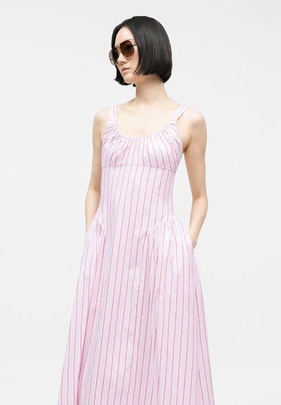 Žena s krátkymi čiernymi vlasmi, ktorá nosí veľké slnečné okuliare a bezrukávové ružovo-bielo pruhované šaty s vreckami.