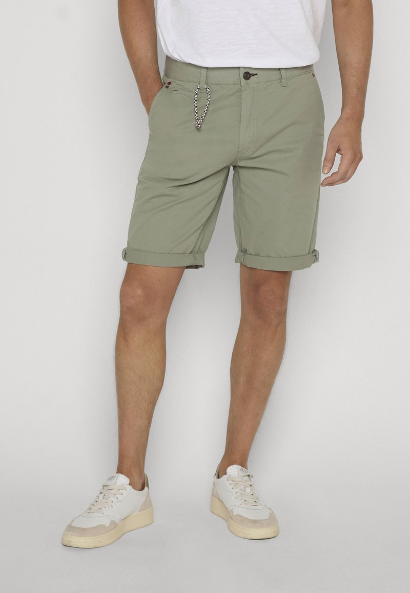 Homme portant un short vert clair retroussé, des baskets blanches et un t-shirt blanc, debout devant un fond uni.