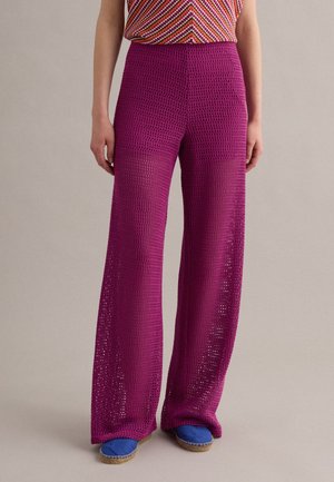 Person trägt hoch taillierte magentafarbene, weit geschnittene Strickhosen, gemustertes ärmelloses Top und blaue Espadrilles vor einfarbigem Hintergrund.