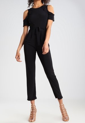 Kvinde iført en sort jumpsuit med kolde skuldre, talje bånd og nøgenfarvede hæle med stropper, står foran en ensfarvet baggrund.