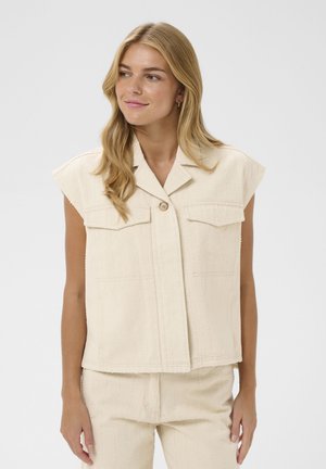 Heledate juustega naine, kes kannab varrukateta kreemikat tekstuuriga nööpidega särk ja sobivaid pükse, vaatab kergelt vasakule neutraalse ilmega.