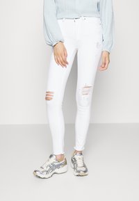 Jeans brancos justos com rasgos desgastados nos joelhos, combinados com uma blusa azul clara e ténis desportivos prateados. Textura suave e ajustada.