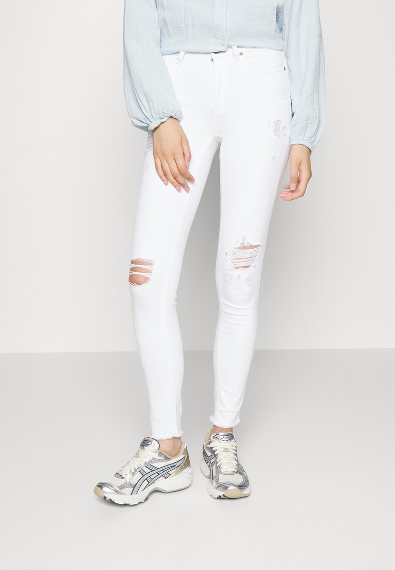 Jeans brancos justos com rasgos desgastados nos joelhos, combinados com uma blusa azul clara e ténis desportivos prateados. Textura suave e ajustada.