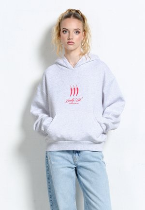 Młoda kobieta o blond włosach w szarej bluzie z czerwonymi papryczkami chili i napisem „Locally Hot”, ubrana w jasnoniebieskie dżinsy, stoi na tle białego tła.