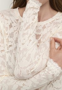 Lace långärmad topp i ljus kräm med intrikata blommönster och vågiga manschetter, med en mjuk, texturerad yta.