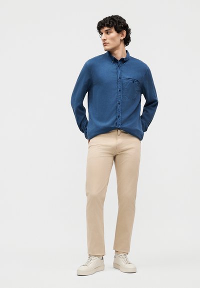 Νέος άνδρας στέκεται με τα χέρια πίσω από την πλάτη του, φορώντας μπλε πουκάμισο με κουμπιά, μπεζ παντελόνι και λευκά αθλητικά παπούτσια, μπροστά σε απλό φόντο.