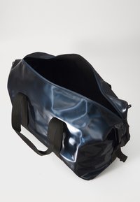 Borsa da viaggio in blu navy lucido con manici neri, aperta per rivelare un interno scuro. Il nome del marchio è elegantemente impresso di lato.