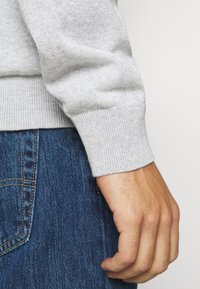 Grauer gestrickter Pullover mit gerippten Bündchen, kombiniert mit blauen Denim-Jeans. Die Textur ist weich und die Passform locker.