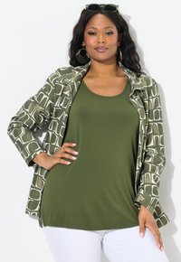 Olivově zelený top bez rukávů pod vzorovanou dlouhou bundou v zelené a bílé barvě s geometrickými vzory. Bílé kalhoty, oversized kruhové náušnice.