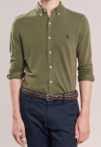 Camicia verde oliva con bottoni, maniche lunghe, colletto e bottoni bianchi. Abbinata a una cintura intrecciata di pelle marrone e pantaloni blu scuro.