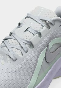 Κοντινό πλάνο ενός ανοιχτόγκρι παπουτσιού Nike για τρέξιμο με αεριζόμενο πλέγμα, γκρι κορδόνια και κυρτό διακοσμητικό σχέδιο κατά μήκος της πλευράς.