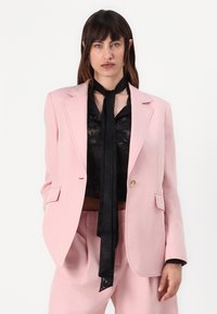 Blazer rosa chiaro in tessuto liscio con spalle strutturate, rever a scollo a V e chiusura con un bottone, abbinato a un top nero trasparente e a una sciarpa.