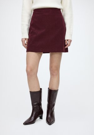 Femme portant une mini-jupe en velours côtelé bordeaux, des bottes en cuir marron foncé mi-mollet et un pull crème à manches longues, debout sur un fond blanc uni.