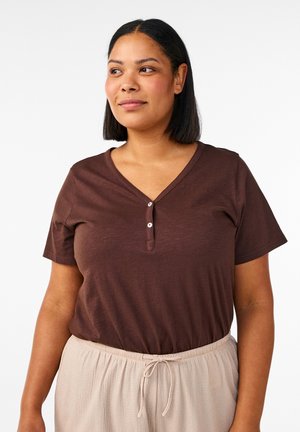 Kvinde med skulderlangt mørkt hår iført en brun kortærmet henley-shirt og beige bukser med snoretræk, står og kigger til siden.