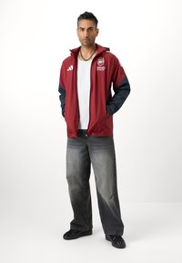 Röd och marinblå zip-up jacka med logotyp, bärs över en vit skjorta, ihop med grå vida jeans och svarta skor. Fristående figur på en enkel bakgrund.