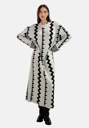 Mujer de pie con las manos en las caderas, vistiendo un vestido largo gris de punto con patrones en zigzag negros y botas negras.