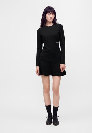 Femme aux longs cheveux noirs portant un haut noir à manches longues, une jupe courte noire, des chaussettes noires et des chaussures noires à lacets sur un fond blanc uni.