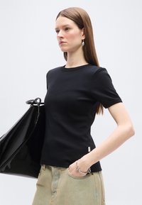 Νεαρή γυναίκα με ίσια καστανά μαλλιά, φοράει μαύρο κοντομάνικο πουκάμισο, μπεζ παντελόνι, ασημένιο βραχιόλι και κρατά μια μεγάλη μαύρη τσάντα στον έναν ώμο.