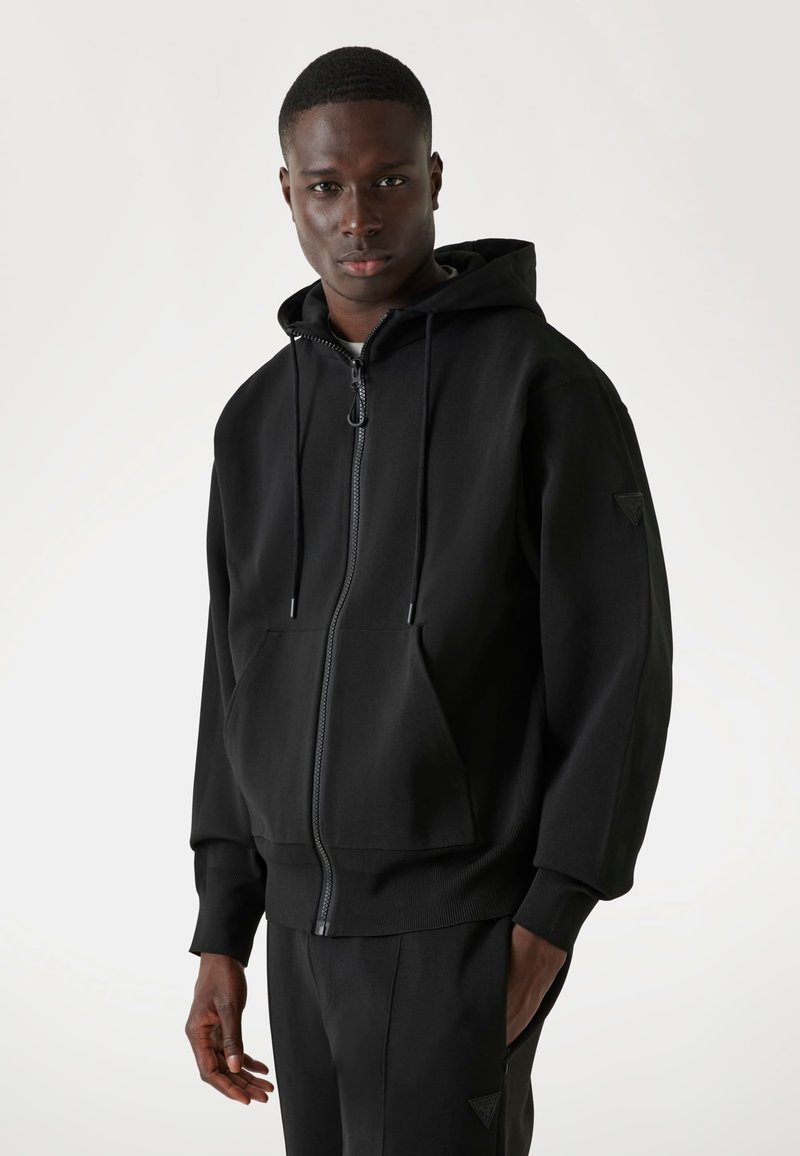 Sudadera con capucha negra hecha de tela suave, presenta una cremallera frontal, cordones, bolsillo tipo canguro y puños de canalé.