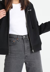 Sudadera negra con cremallera sobre una camiseta blanca, combinada con jeans gris de cintura alta. La sudadera cuenta con cordón y un sutil detalle del logo.