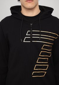 Black zip-up hoodie with a smooth texture and a gold graphic design featuring parallel lines on the front. Includes a hood and drawstrings.