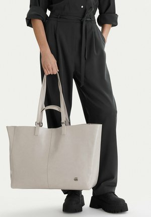 Persona vestida con traje negro sostiene un gran bolso tote beige con asas dobles, de pie contra un fondo claro y liso.