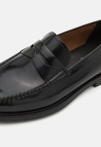 Mocassin penny en cuir noir poli avec coutures moc toe et doublure en cuir sur fond blanc.