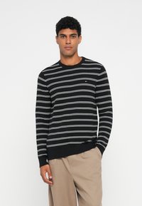 Maglione nero a maglia con strisce orizzontali bianche, scollo rotondo e polsini a coste. Indossato da un modello con pantaloni di colore neutro.